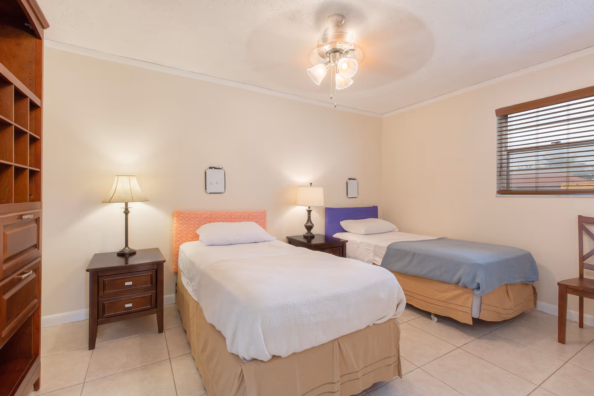 A bedroom with two single beds, each with a headboard in different colors (orange and purple). Both beds have white bedding and beige bed skirts. There are two wooden nightstands, each with a lamp, positioned between and beside the beds. A wooden chair is placed near a window with wooden blinds. The room has beige walls, tiled floors, and a ceiling fan with lights.