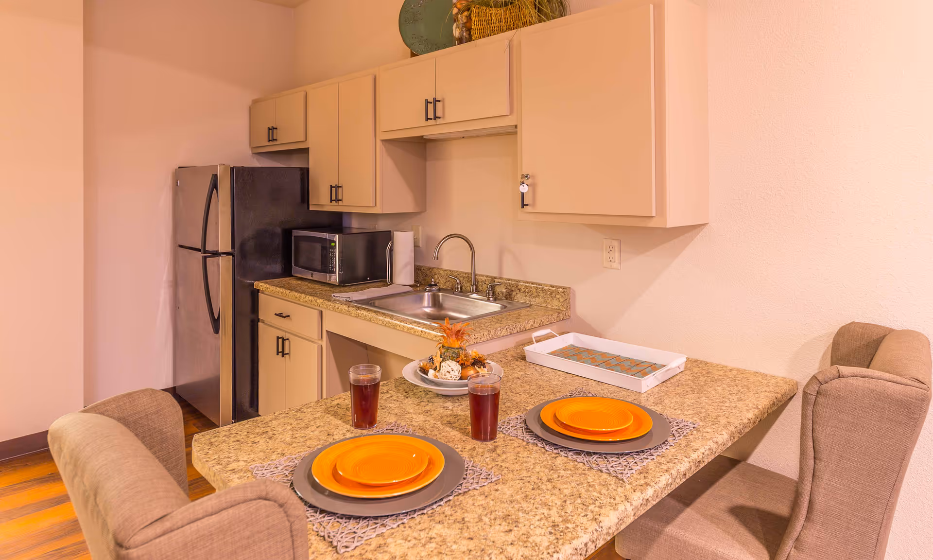 A small kitchen area with beige cabinets, a stainless steel refrigerator, a microwave, and a sink. In front of the kitchen counter is a dining area with two place settings, each with an orange plate on a gray placemat and a glass of dark beverage. Two cushioned chairs are positioned at the counter. The countertop has a decorative centerpiece and a white tray.