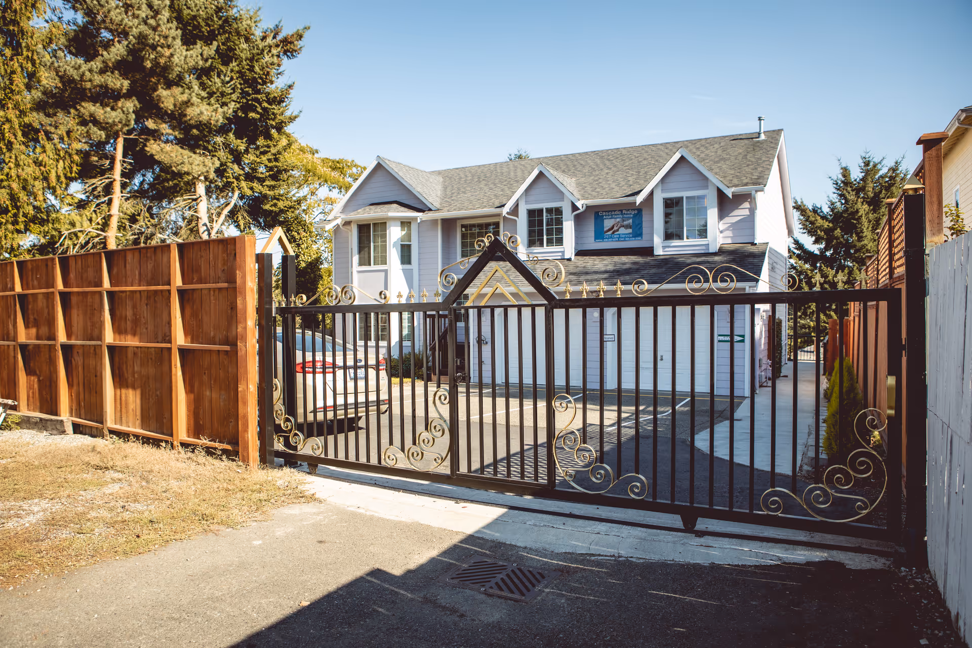 View of a two-story residential building behind a decorative black metal gate with gold accents. The building has light gray siding, multiple windows, and a sign on the upper front wall. There is a car parked inside the gated driveway, and tall trees are visible in the background under a clear blue sky.