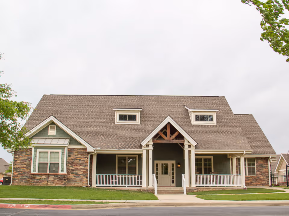 Front exterior view of a single-story residential building with a large gabled roof, stone and siding facade, a covered porch with white railings, and a central entrance door. The building is surrounded by a well-maintained lawn and trees.