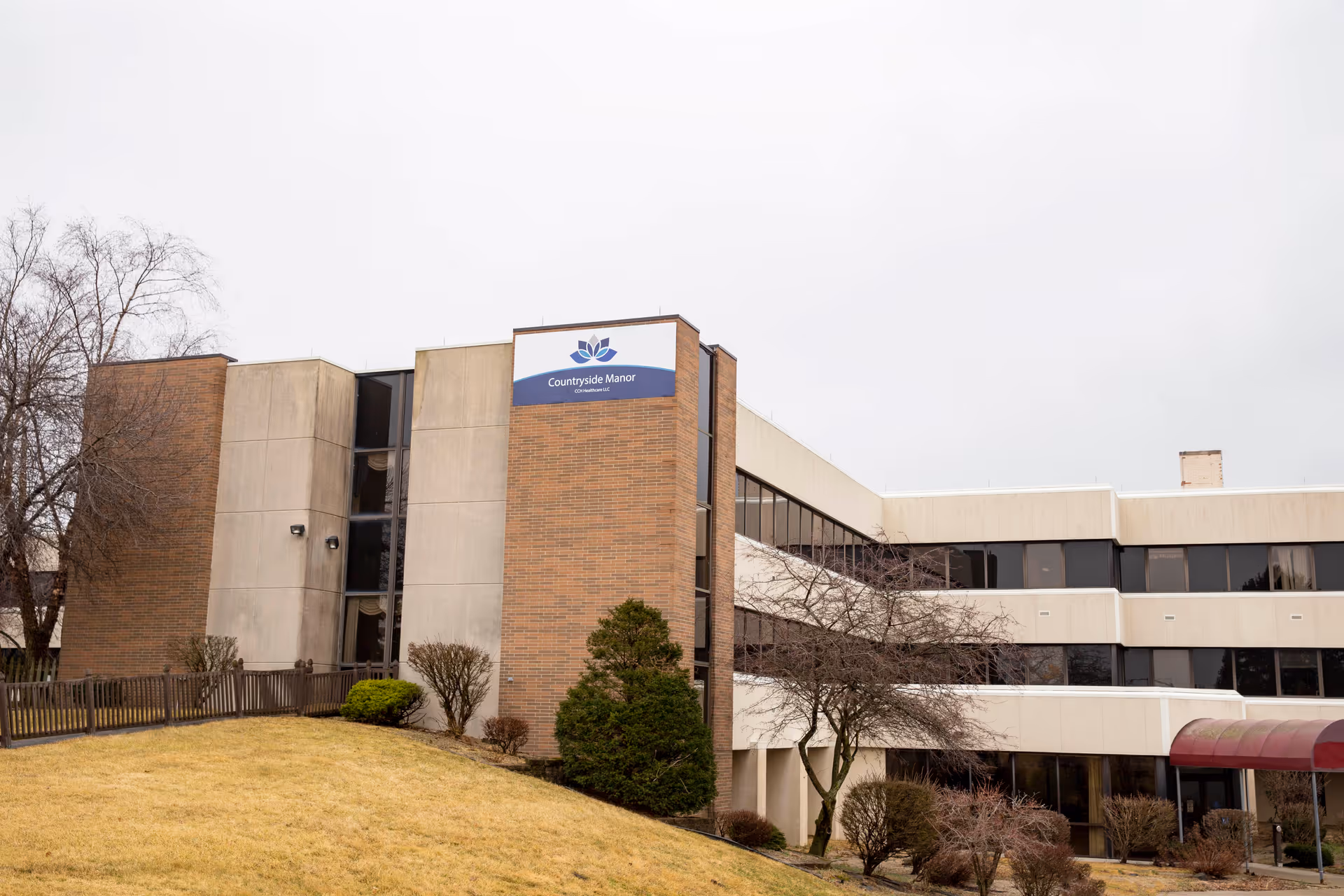 Exterior view of a multi-story building with a brick and concrete facade, featuring large windows and a sign that reads 'Countryside Manor Healthcare'. There is a grassy area with bushes and trees in front of the building under an overcast sky.