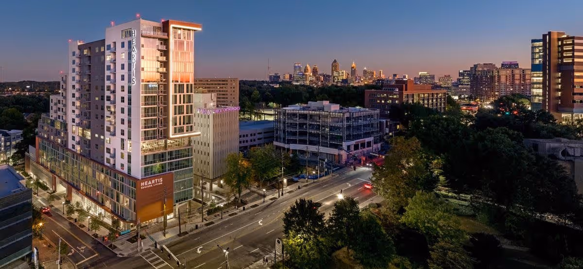 Aerial evening view of the Heartis Buckhead Senior Living building in an urban area with city skyline in the background, surrounded by streets, trees, and other buildings.