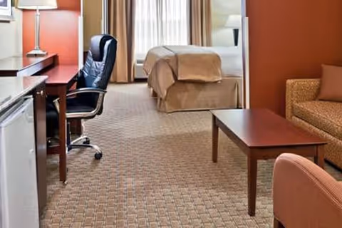 Interior view of a senior living facility room featuring a bed with beige bedding near a window with curtains, a wooden desk with a black office chair, a small refrigerator, a wooden coffee table, a patterned sofa, and an upholstered armchair on a carpeted floor.