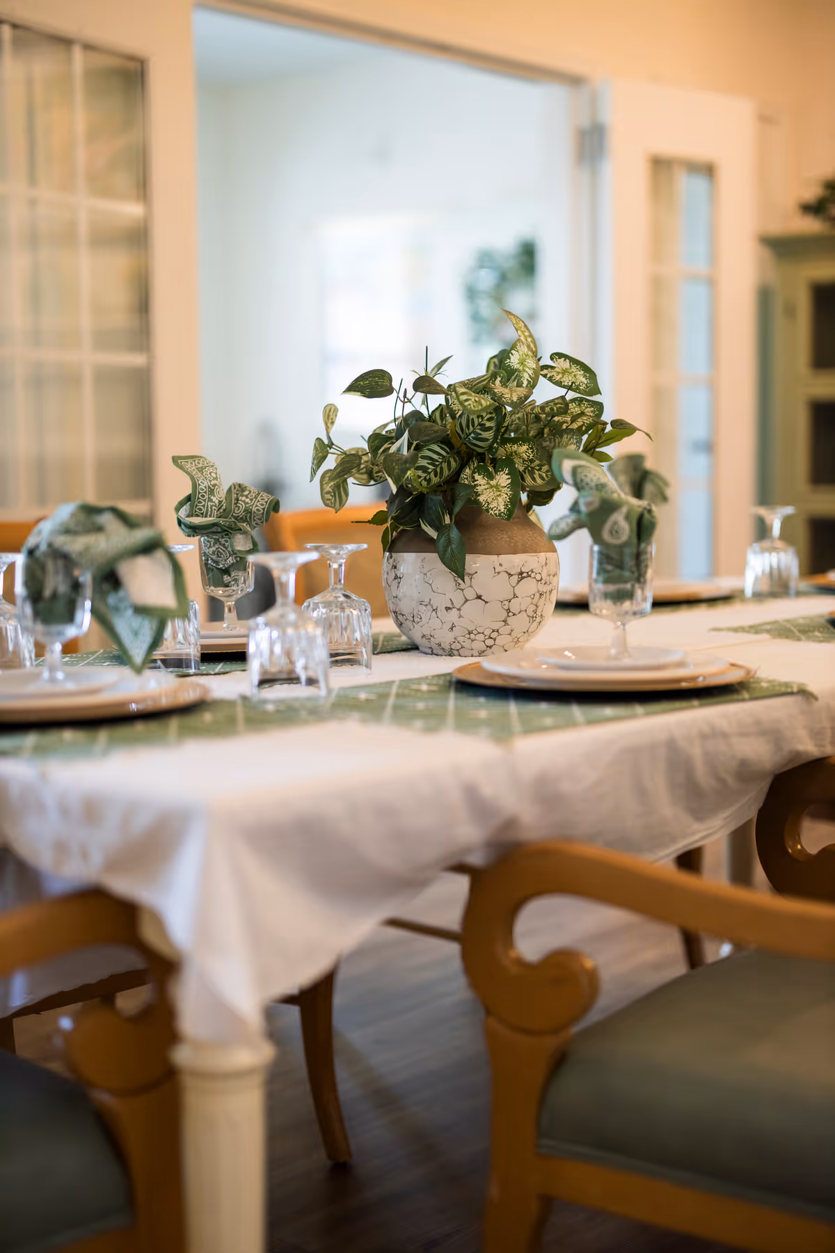 A dining table set with plates, glassware, green patterned napkins, and a potted plant centerpiece in a bright dining room.