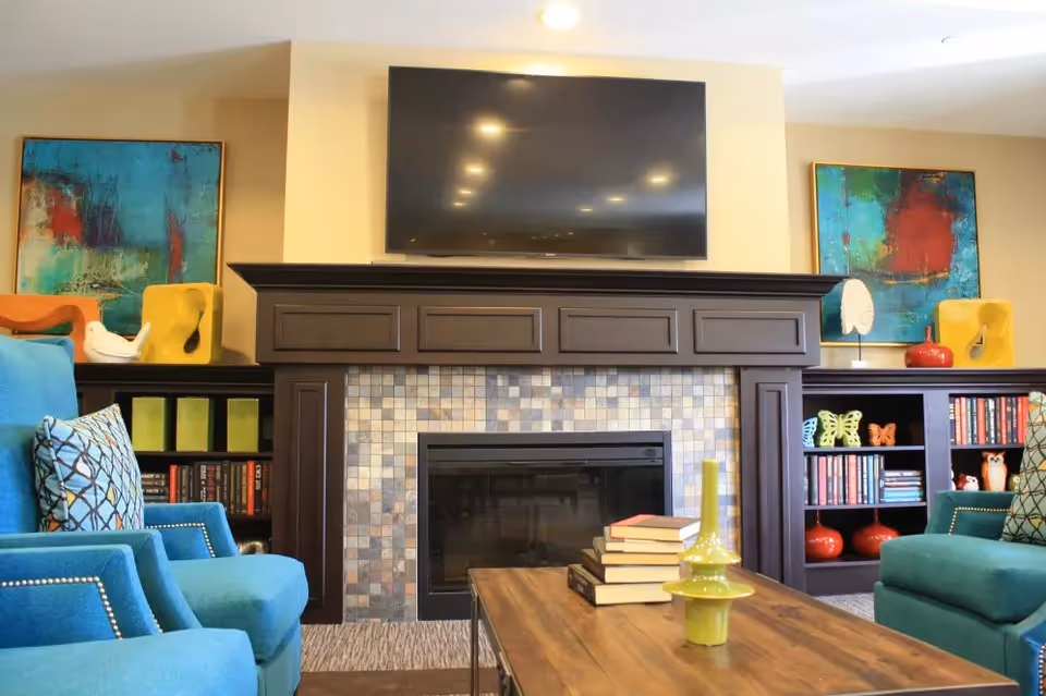 Living room with a tiled fireplace and wall-mounted TV, turquoise armchairs, bookshelves and a wooden coffee table with stacked books and decorative items.