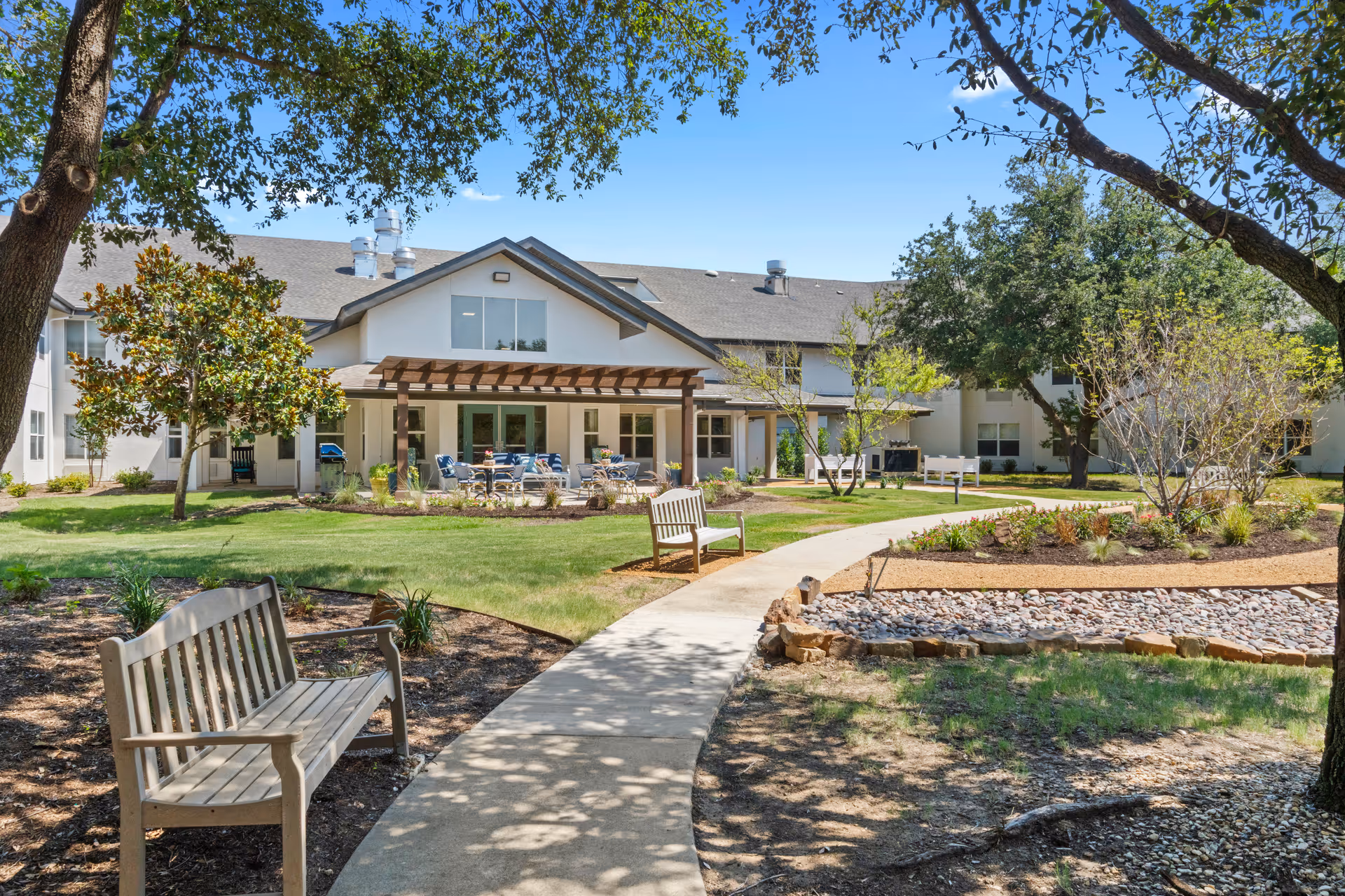 Outdoor garden area at The Grove on Forest Lane featuring a paved walkway, wooden benches, landscaped plants, trees, and a two-story building with a covered patio and outdoor seating under a pergola.