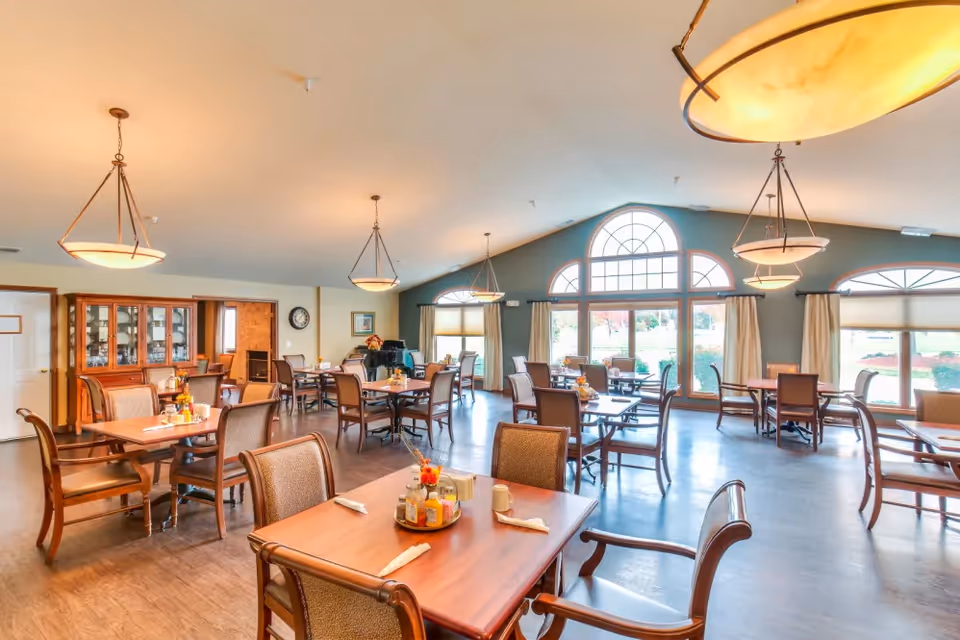 Bright, spacious dining room with multiple wooden tables and chairs, large arched windows, and hanging pendant lights.