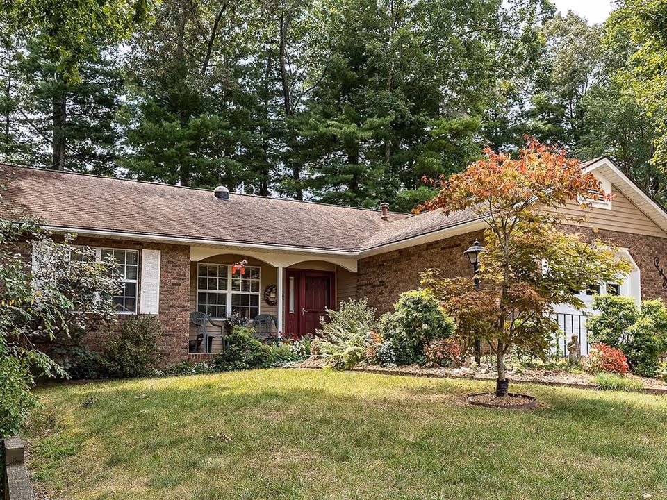 Front exterior view of a single-story brick house with a brown shingled roof, a red front door, white window shutters, and a small porch with two chairs. The house is surrounded by green grass, bushes, and trees, with a small tree with red leaves in the front yard.