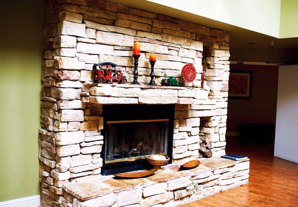 Stone fireplace with a mantle decorated with candles, miniature vehicles and bowls, set in a room with hardwood floors.