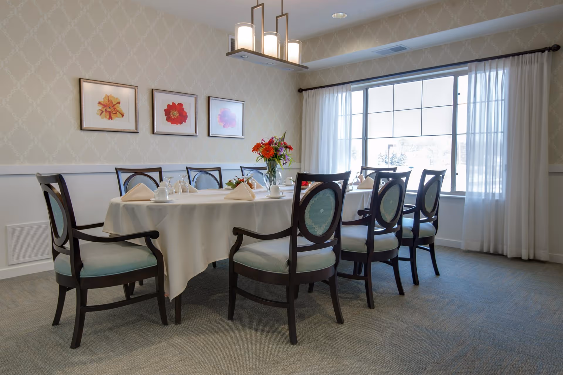 A dining room with a large rectangular table covered with a white tablecloth, set with napkins, cups, and a vase of colorful flowers. The room has eight wooden chairs with light blue cushioned seats and backs. There are three framed floral artworks on the wall and a large window with white curtains letting in natural light.