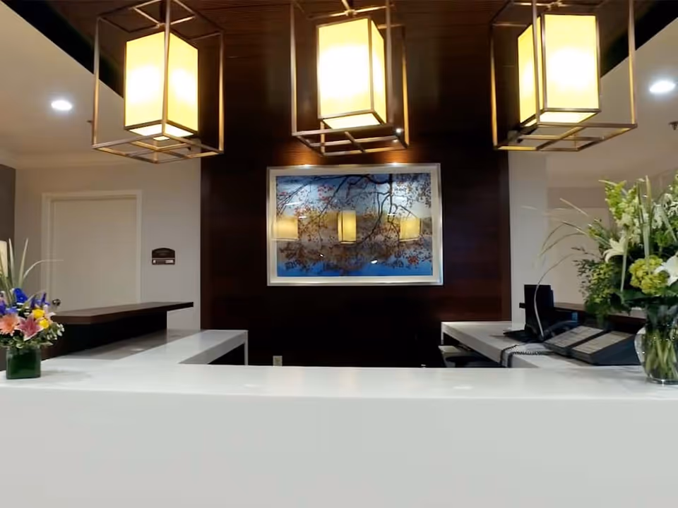Reception desk area with a white countertop, two vases of flowers on either side, three large rectangular pendant lights hanging from the ceiling, a framed picture of tree branches with flowers on a dark wood-paneled wall, and office equipment including a phone and computer.