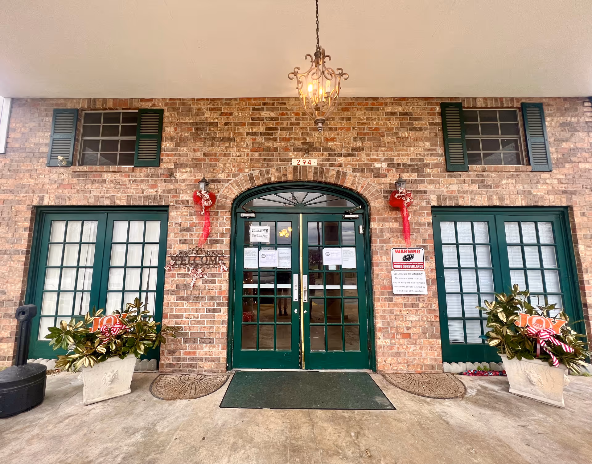 Front entrance of Rayville Nursing & Rehabilitation Center, Inc. featuring double green glass doors with brick walls, two large green-framed windows on each side, decorative plants in white pots, holiday decorations, and a hanging lantern light above the entrance.