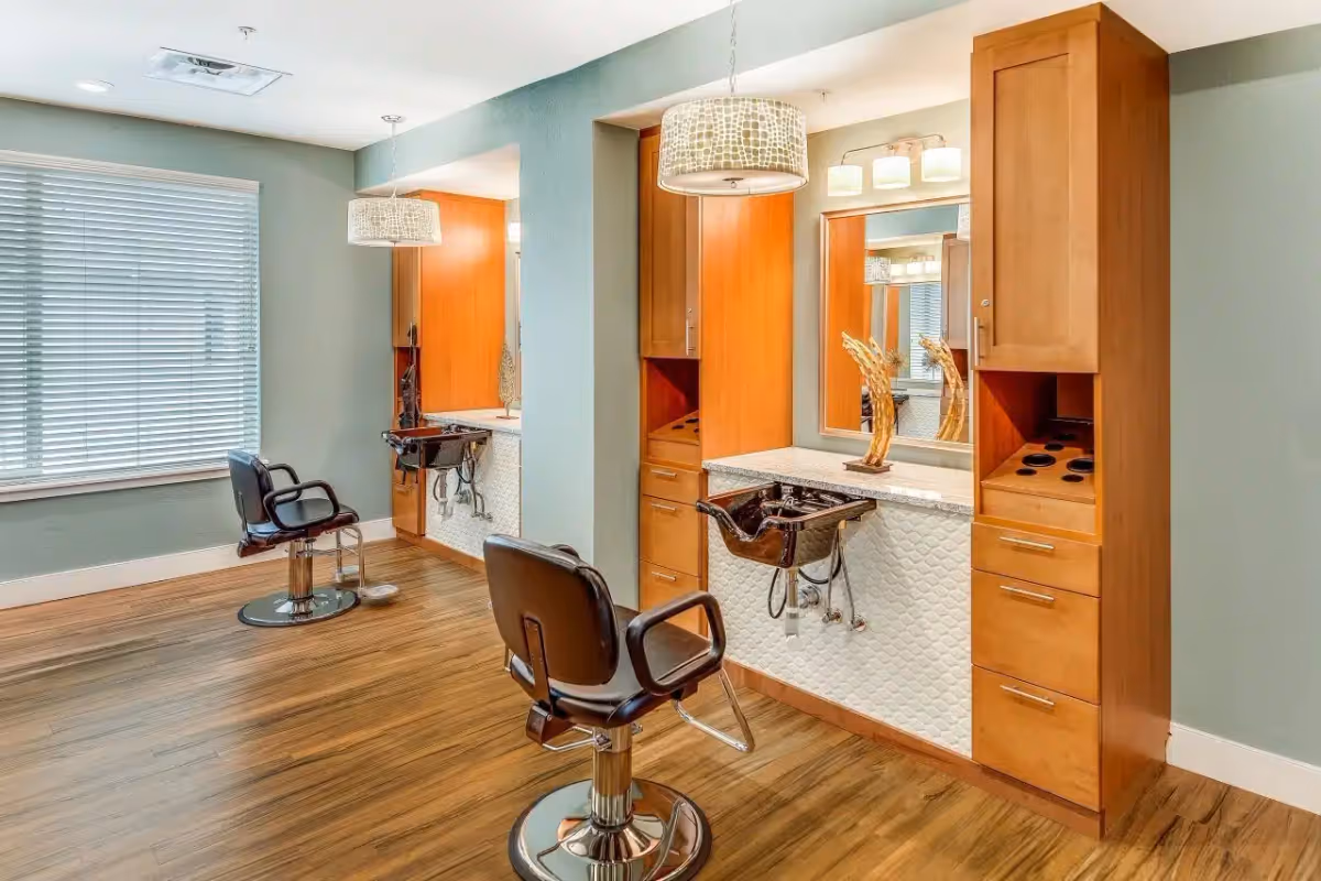 Interior view of a hair salon area with two black salon chairs in front of two wooden stations with mirrors, sinks, and pendant lights. The floor is wooden, and there is a window with blinds on the left wall.