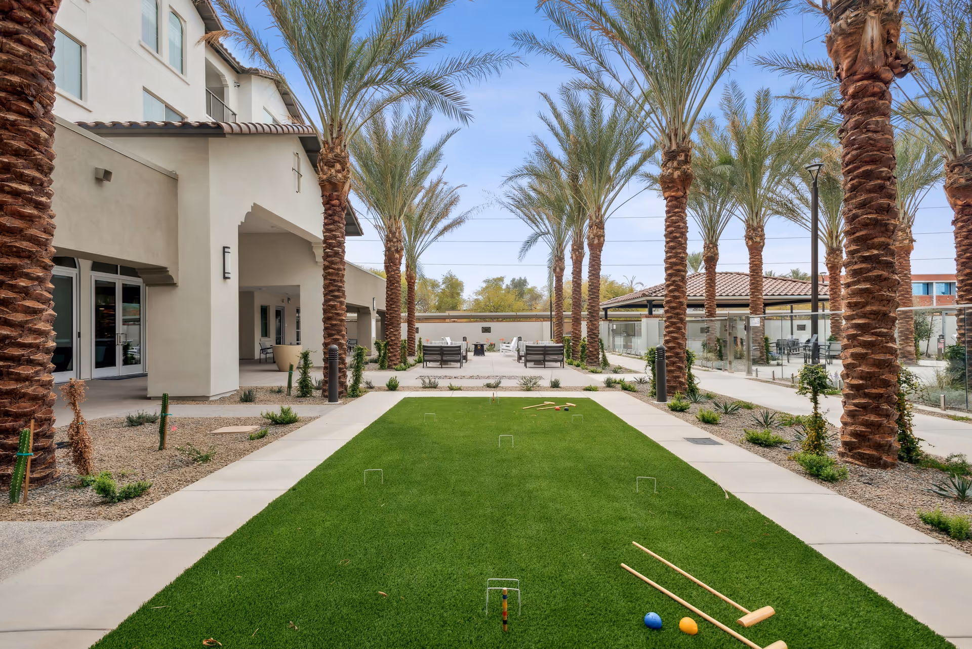 Outdoor courtyard area at Overture North Scottsdale featuring a green lawn with croquet equipment set up, surrounded by palm trees and walkways. There are seating areas with chairs and tables in the background near a building with a tiled roof and glass fencing.