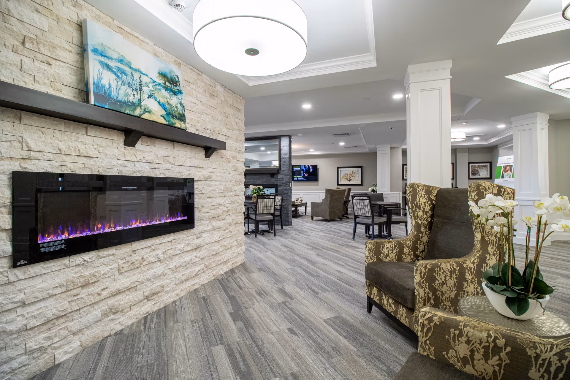 A spacious and well-lit living room area in a senior living facility featuring a modern electric fireplace mounted on a stone wall with a painting above it. The room has comfortable upholstered chairs, tables, decorative plants, and framed artwork on the walls. The flooring is wood-patterned, and the ceiling has recessed lighting and large circular light fixtures.
