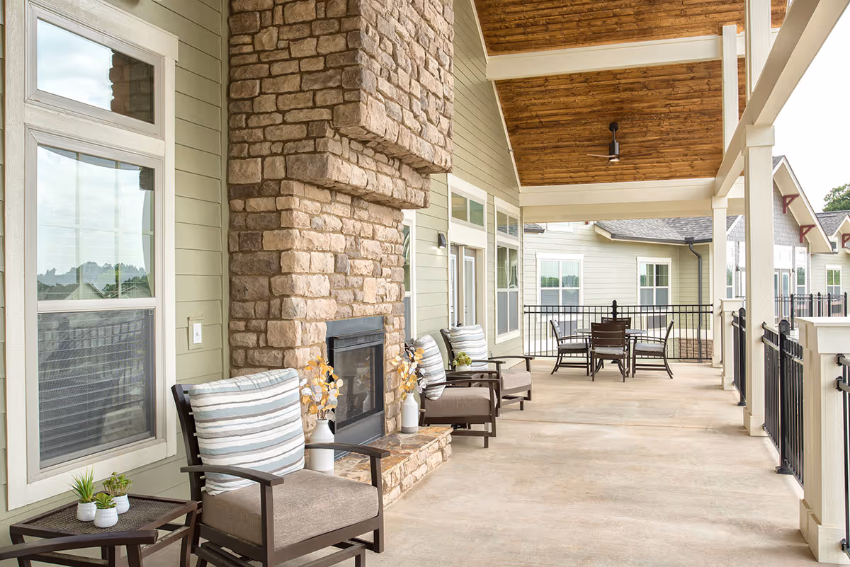 Covered outdoor patio area with a stone fireplace, cushioned chairs with striped pillows, small side tables with potted plants, and a dining table with chairs in the background. The patio has a wooden ceiling with a ceiling fan and is surrounded by a black metal railing.