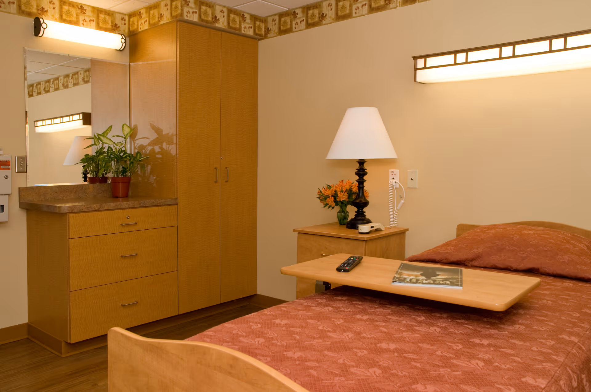 A senior living facility bedroom with a single bed covered in a reddish-brown patterned bedspread. There is a wooden overbed table with a remote control and a magazine on it. Next to the bed is a wooden nightstand with a black lamp, a small vase of orange flowers, and a wall-mounted telephone. On the left side of the room, there is a wooden dresser with three drawers and a countertop with two potted plants. Above the dresser is a large mirror and a wall light. The walls are beige with a decorative border near the ceiling.