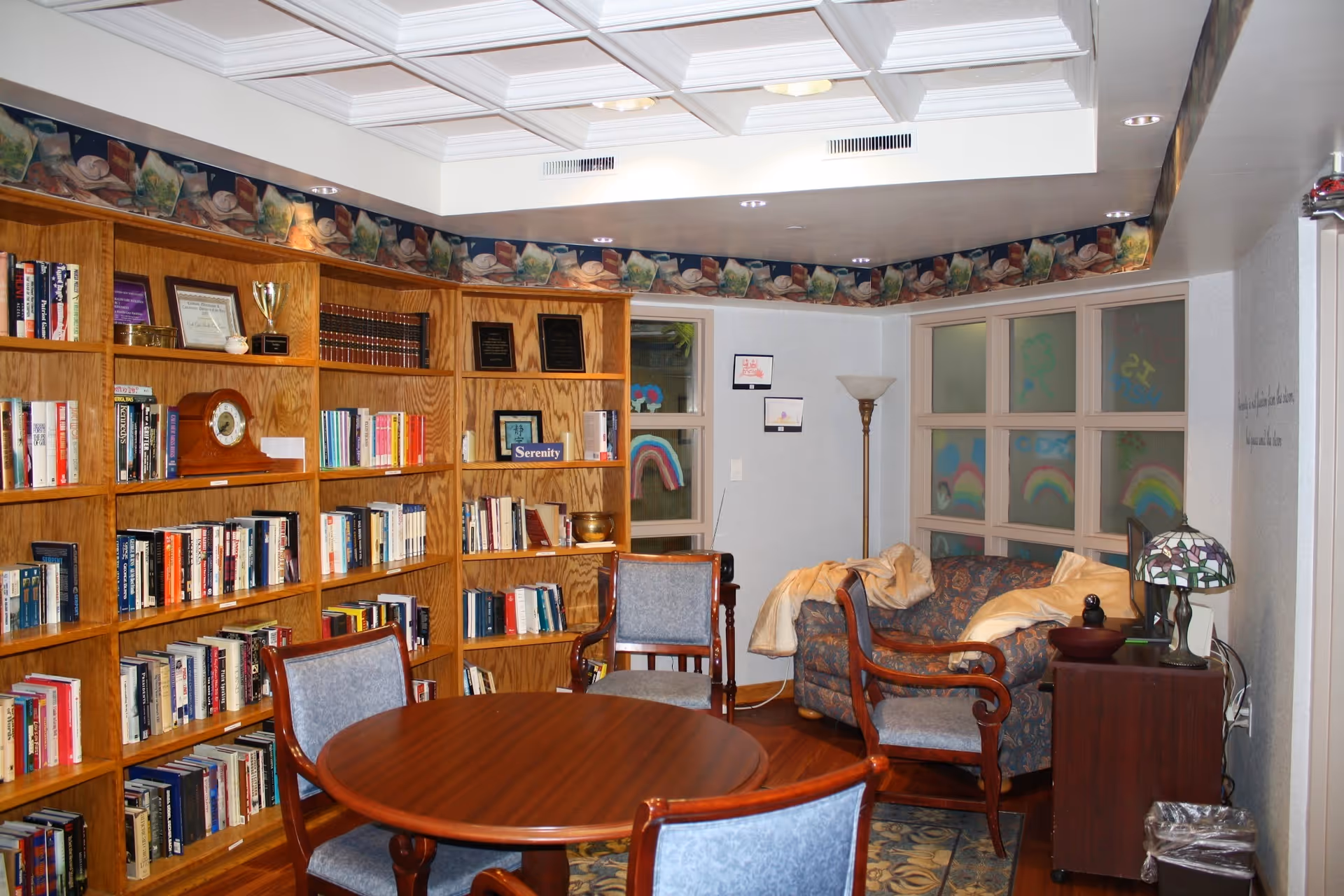 A cozy room with wooden bookshelves filled with books and decorative items, a round wooden table surrounded by four chairs with blue upholstery, a patterned couch with a blanket, a floor lamp, and a small cabinet with a stained glass lamp. The walls have windows decorated with colorful rainbow drawings.