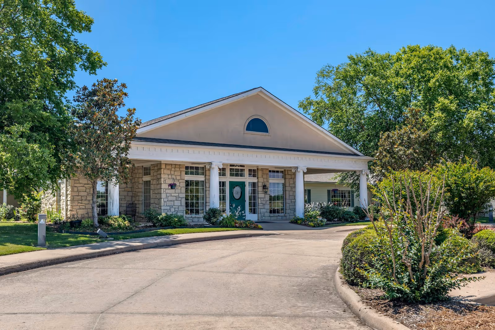 Front exterior view of a single-story building with stone and beige siding, white columns, a green door, and large windows surrounded by well-maintained landscaping and trees under a clear blue sky.