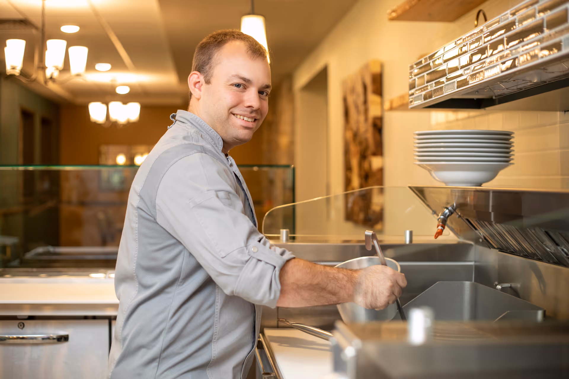 A man in a gray shirt smiling while preparing food in a kitchen area with plates stacked on a shelf above a stainless steel counter.
