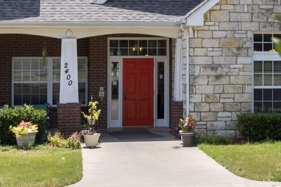 Front entrance of a senior living facility with a red door, brick and stone exterior walls, potted plants, and the number 2400 displayed on a white column.