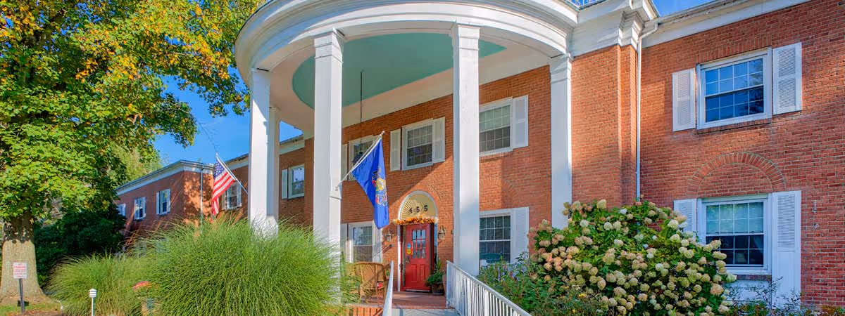 Front exterior view of a red brick building with white columns and a covered entrance. There are two flags displayed near the entrance, an American flag and a blue flag. The building has white window shutters and is surrounded by green bushes and trees under a clear blue sky.