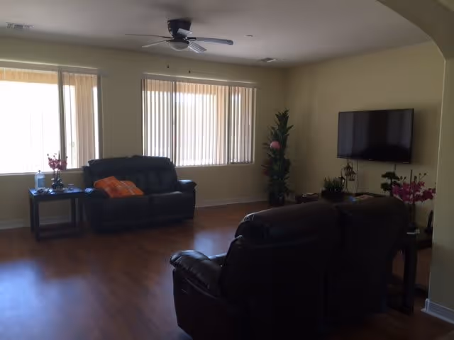 Sunlit living room with leather recliners, a wall-mounted TV, ceiling fan, and large windows with vertical blinds.