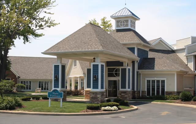 Exterior view of Sprenger Health Care Anchor Lodge Retirement Village showing the main entrance with a covered drop-off area, landscaped greenery, and a clear blue sky.