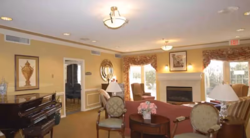 Cozy common room with sofas, armchairs, a fireplace, piano, and large windows.