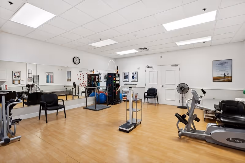 A fitness room in a senior living facility with exercise equipment including a stationary bike and a recumbent bike, a scale in the center, chairs along the walls, a large mirror on one side, and various exercise balls and equipment stored on shelves. The room has wood flooring, white walls, and fluorescent ceiling lights.
