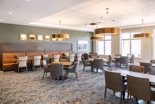 A spacious dining room with multiple round and rectangular tables surrounded by chairs. The room features large windows allowing natural light, modern pendant lights hanging from the ceiling, and a cushioned bench along one wall with framed artwork above it. The carpet has a patterned design and the walls are painted in neutral tones.