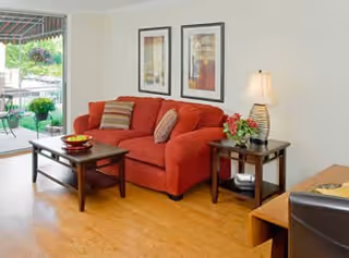 Sunlit living room with an orange sofa, coffee table, side table with a lamp, framed artwork, and a sliding glass door opening to a patio.
