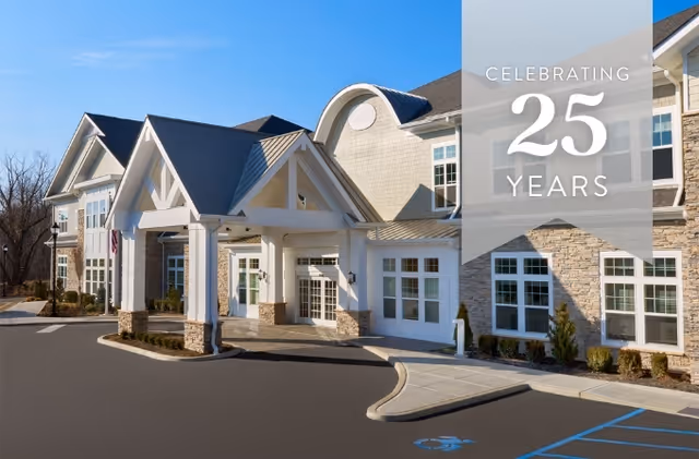 Exterior view of The Bristal Assisted Living at Lake Success building entrance with a covered drop-off area, stone and siding facade, multiple windows, and a clear blue sky. A banner overlay on the right side reads 'Celebrating 25 Years'.