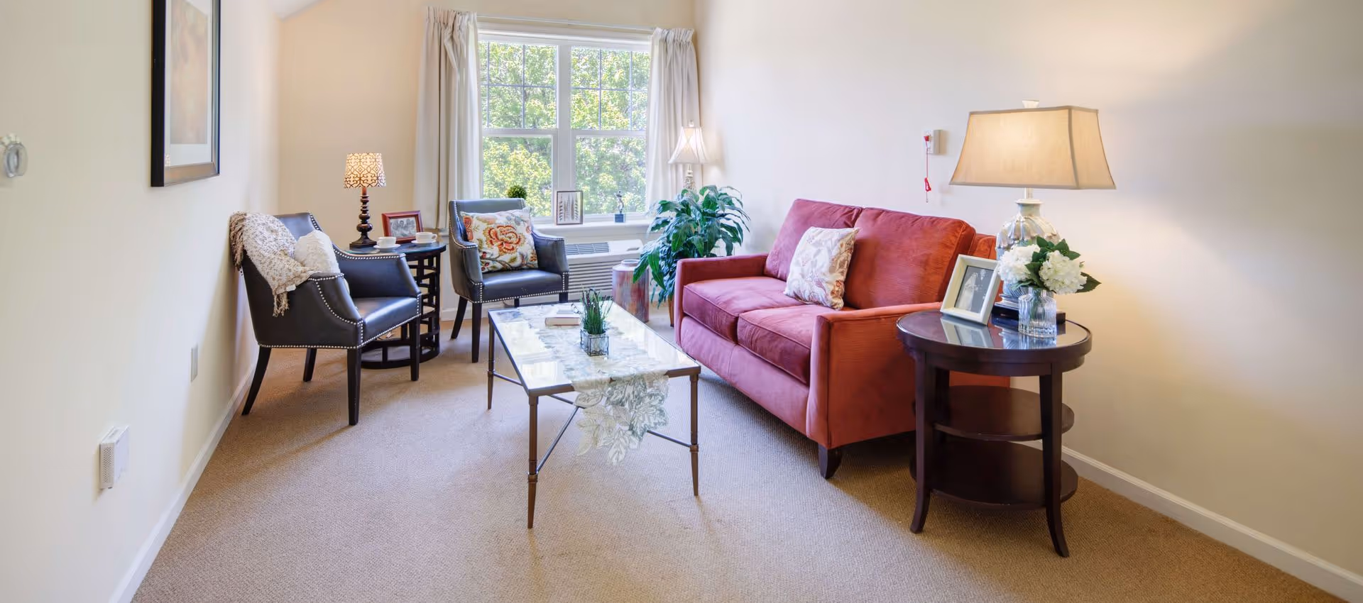 A cozy living room with a red sofa, two dark armchairs with decorative pillows, a glass coffee table with a lace runner, and two side tables with lamps and framed photos. A large window with white curtains lets in natural light, and a green plant is placed near the window.
