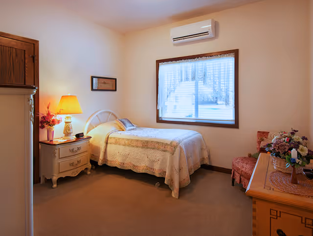 A cozy bedroom with a single bed, nightstand and lamp, window with lace curtains, dresser and chair with floral decor.