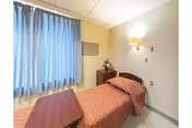 A small, simple bedroom in a rehabilitation and nursing center with a single bed covered in a brown quilted bedspread, a wooden bedside table with a small flower arrangement, a wooden overbed table, beige walls, and a window with blue curtains allowing natural light to enter.