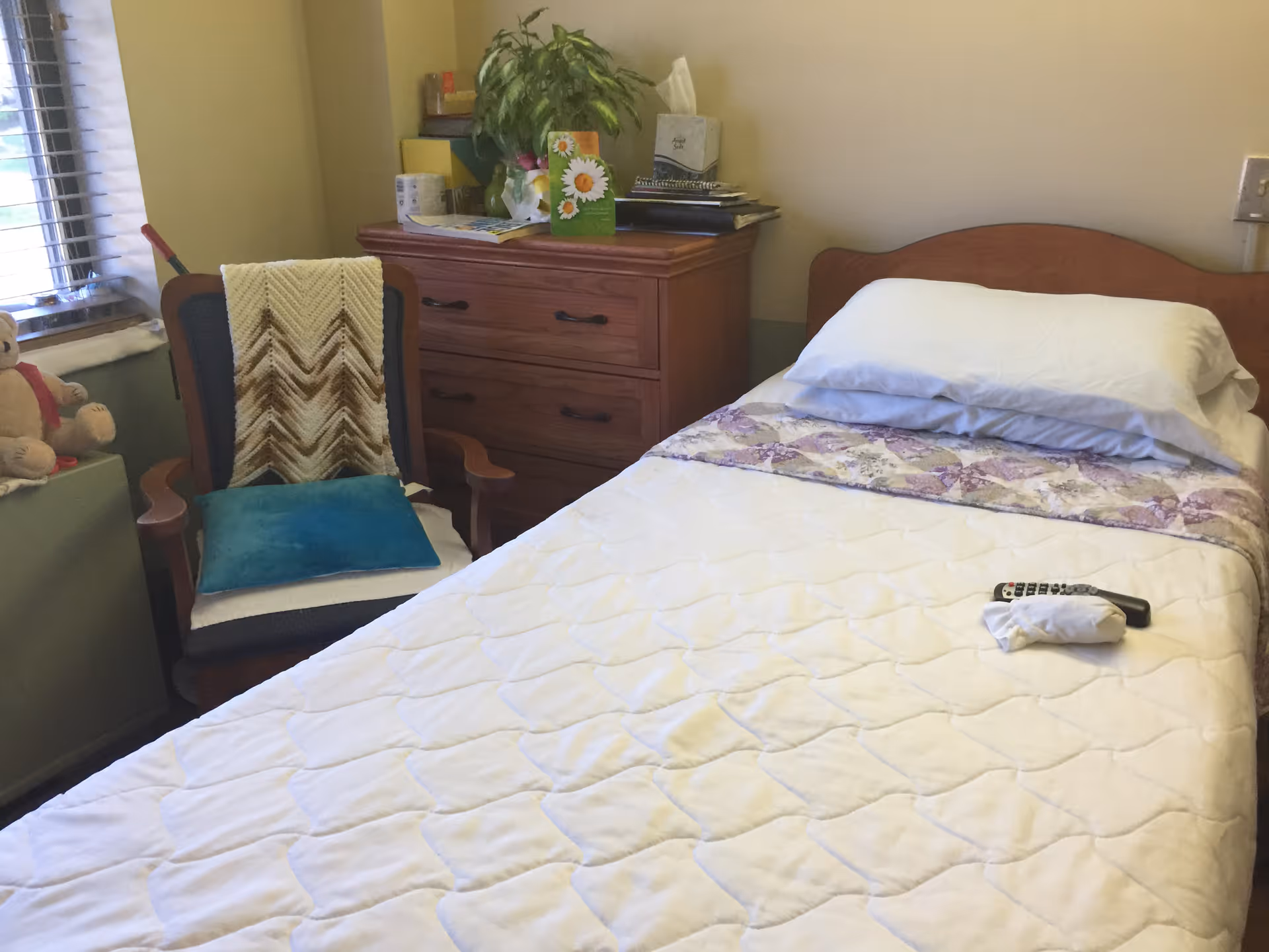 A neatly made single bed with a white quilt and a decorative purple strip near the headboard. A TV remote and a white cloth are placed on the bed. Next to the bed is a wooden dresser with various items on top including a plant, a tissue box, and some papers. To the left of the dresser is a cushioned chair with a blue pillow and a knitted throw draped over the back. A window with blinds and a teddy bear on the windowsill are visible in the background.