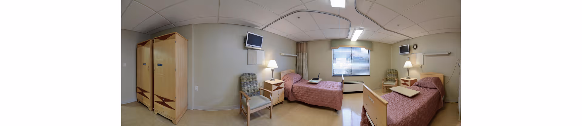Shared nursing facility bedroom with two single beds, bedside tables and lamps, chairs, wardrobes, wall-mounted TVs, and a window.