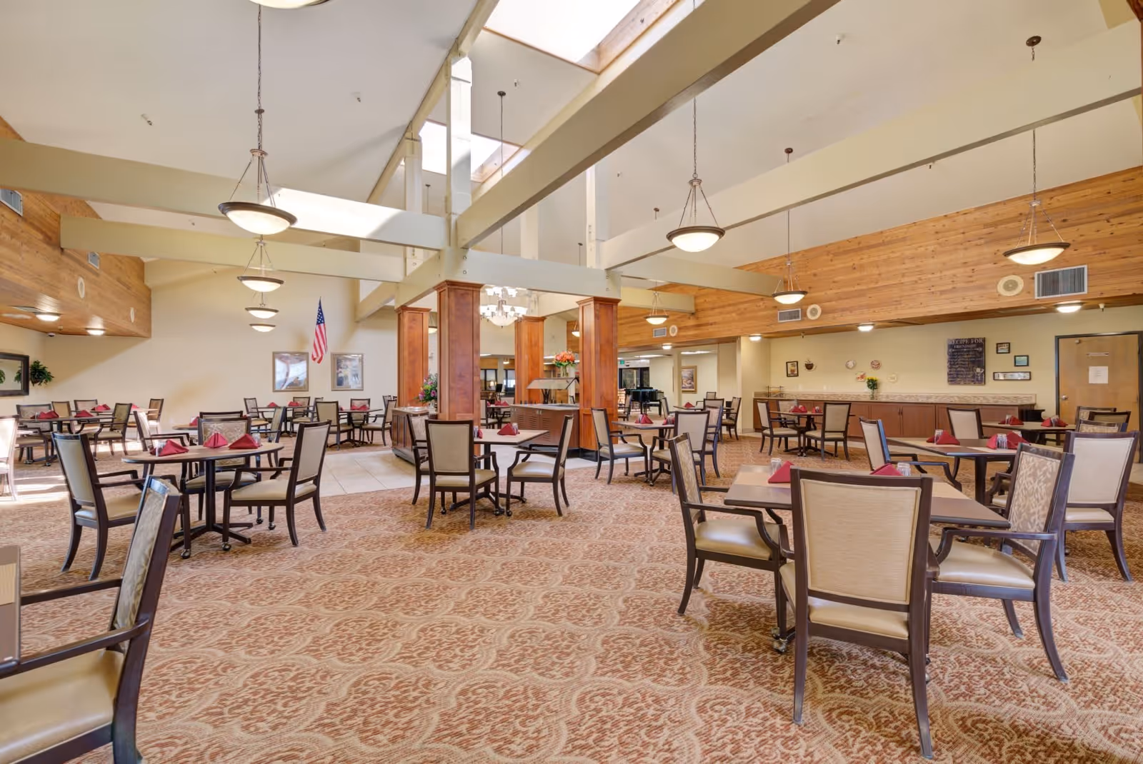 Spacious dining room with multiple tables and chairs arranged neatly, each table set with folded red napkins. The room features high ceilings with exposed beams, hanging light fixtures, and large windows allowing natural light to fill the space. The walls are decorated with framed pictures and an American flag is visible in the background.