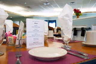 A close-up view of a dining table set with white plates, silverware, glasses with white napkins folded inside, and a menu card placed upright in the center. The background shows a softly lit dining area with additional tables and chairs.