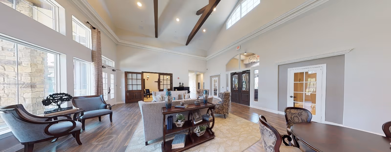 A spacious and well-lit living room area with high vaulted ceilings featuring exposed wooden beams. The room has large windows on one side allowing natural light to fill the space. There are several seating arrangements including a sofa, armchairs, and a small table with chairs. The floor is wooden with a large area rug in the center. Decorative items such as vases and books are placed on a wooden console table behind the sofa. Double doors and an arched doorway lead to other parts of the facility.
