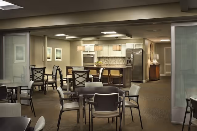 A spacious dining area with multiple tables and chairs arranged neatly. In the background, there is a modern kitchen with white cabinets, a stainless steel refrigerator, an oven, and a kitchen island with bar stools. The room is well-lit with ceiling lights and pendant lamps over the kitchen island. The walls are decorated with framed artwork.