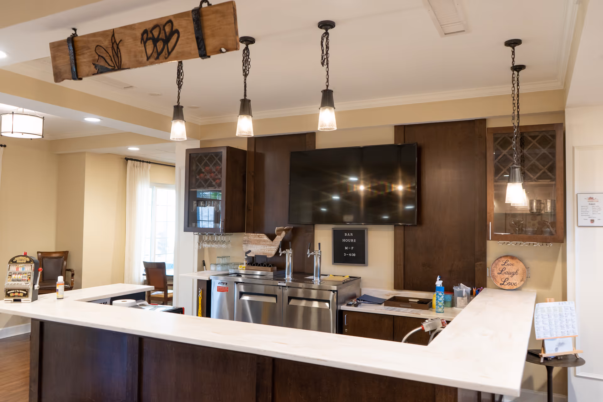 Interior view of a bar area in a senior living facility with a white countertop, dark wood cabinetry, a large flat-screen TV mounted on the wall, and three pendant lights hanging from the ceiling. There are bar taps, glassware, and a sign displaying bar hours. A decorative wooden sign with the words 'Love Laugh Love' is on the counter.