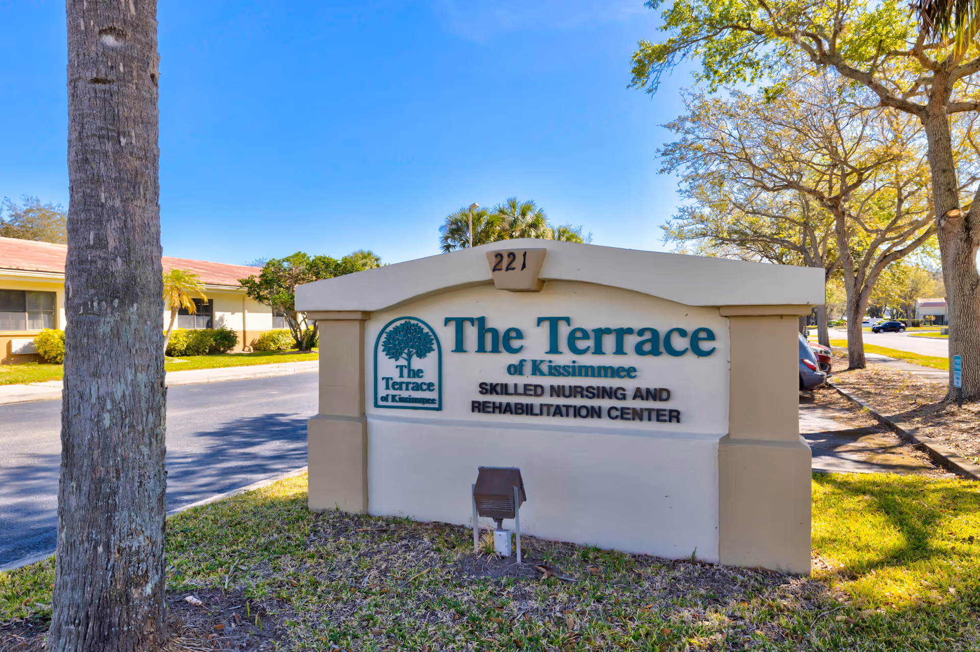 Outdoor view of a sign for The Terrace of Kissimmee Skilled Nursing and Rehabilitation Center, situated on a grassy area near a road with trees and parked cars in the background under a clear blue sky.
