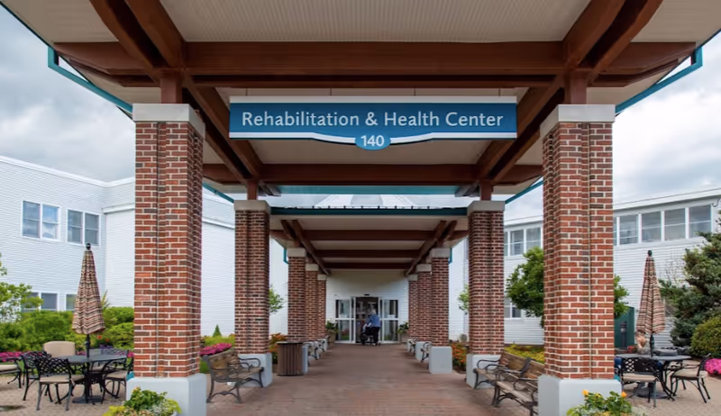 Covered brick-pillared entrance walkway leading to a Rehabilitation & Health Center with benches and outdoor seating.