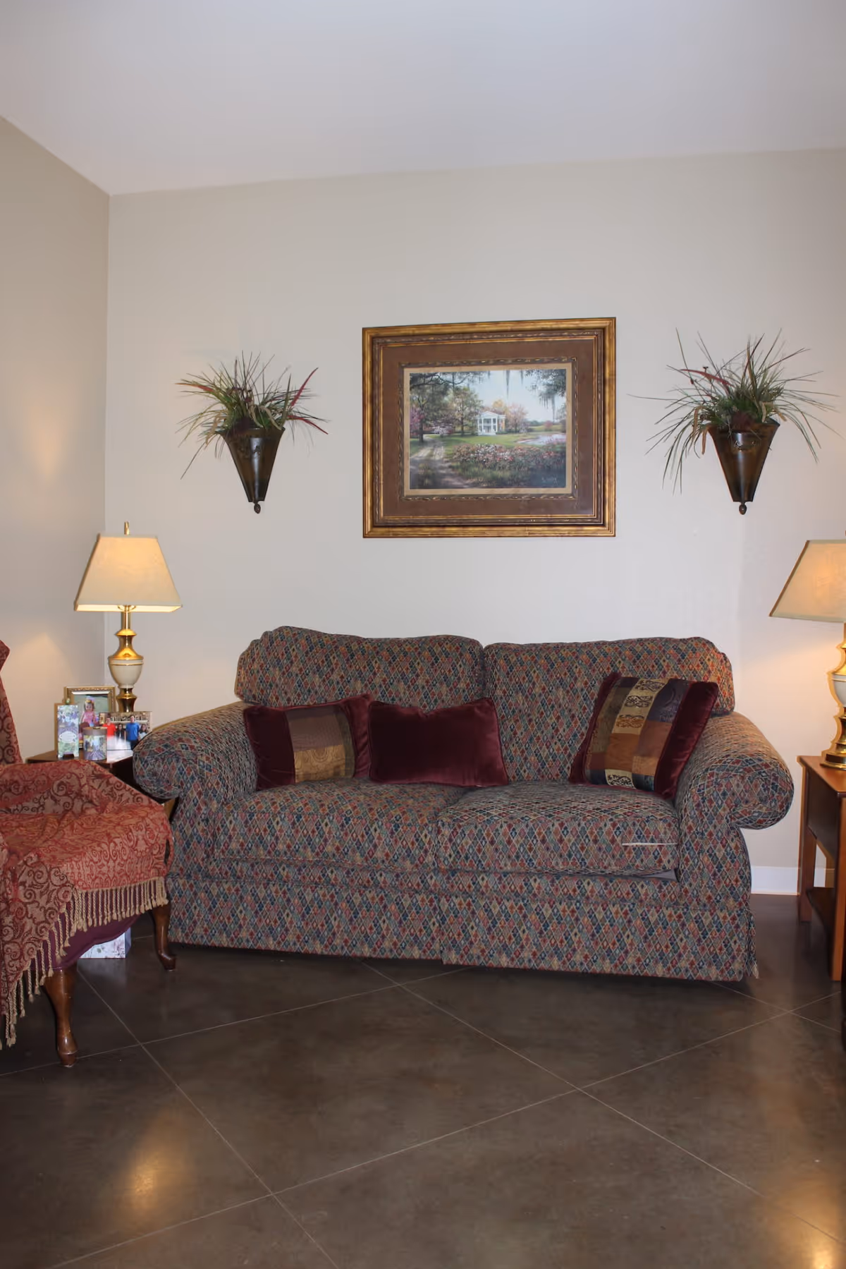 A cozy living room area with a patterned sofa adorned with decorative pillows. On either side of the sofa are wooden side tables with table lamps. Above the sofa hangs a framed painting of a garden scene, flanked by two wall-mounted planters with green plants. To the left, a red upholstered chair with a fringed throw is partially visible.