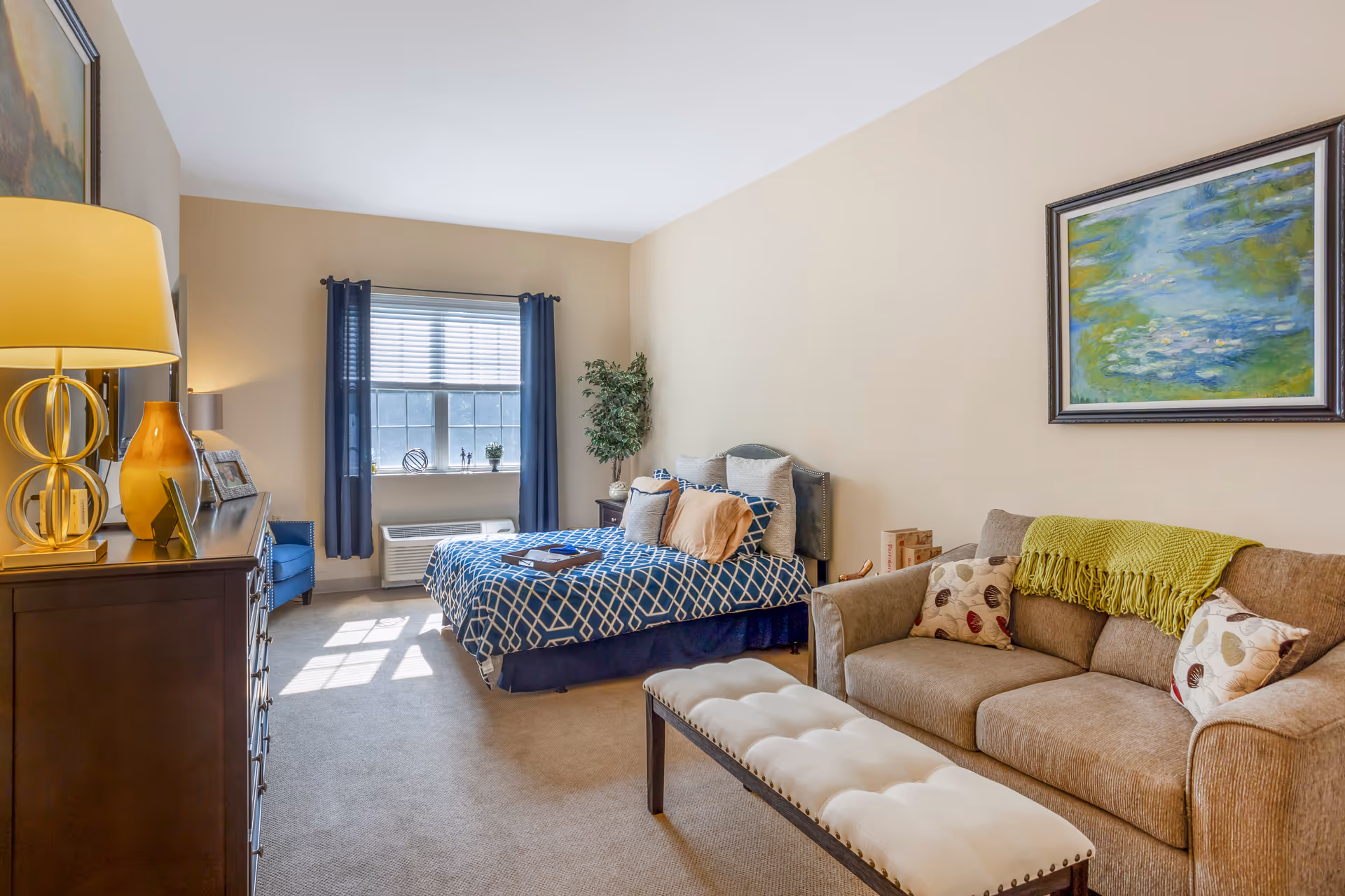 A well-lit bedroom featuring a bed with blue and white patterned bedding, a brown sofa with decorative pillows and a green throw, a cushioned bench, a wooden dresser with a lamp and framed photos, a blue armchair near a window with blue curtains, and a framed painting on the wall.