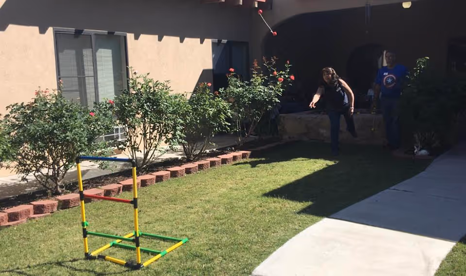 Two people playing a lawn toss game toward a colorful ladder target on a grassy courtyard outside a beige building.