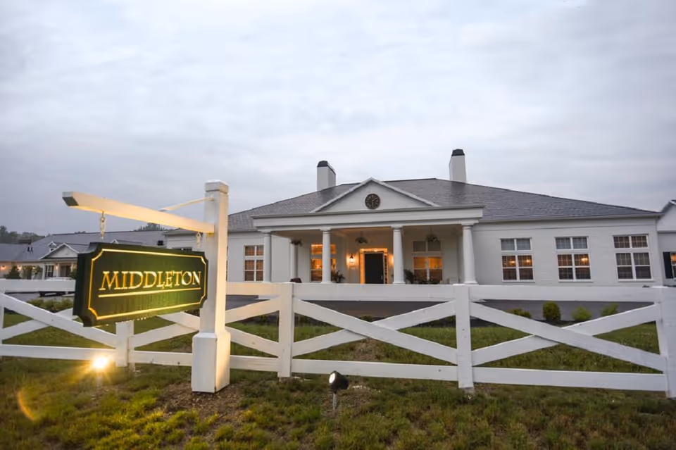 Front exterior of Middleton Senior Living building at dusk with an illuminated 'Middleton' sign and white fence.