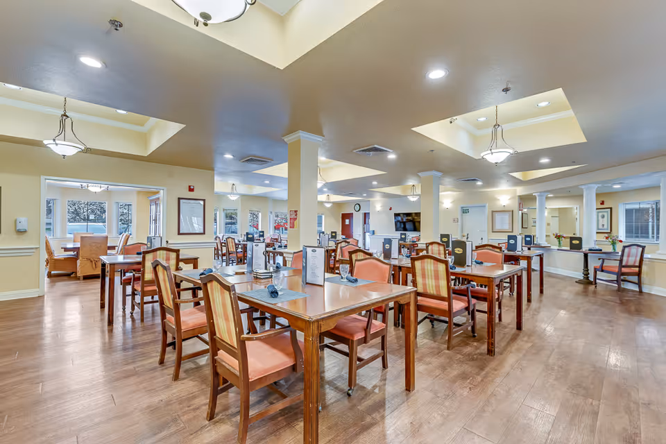 A spacious dining room in a senior living facility with multiple wooden tables and chairs arranged neatly. Each table has place settings including glasses and napkins, and menus or informational cards are placed on the tables. The room has a warm color scheme with beige walls, wooden flooring, and ceiling lights including recessed lighting and hanging fixtures. Large windows allow natural light to enter, and there is a television mounted on the far wall.